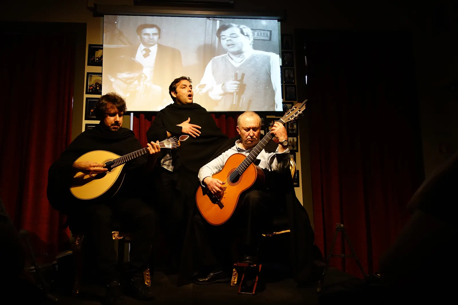 Trzech mężczyzn w czarnych pelerynach akademickich na scenie z czerwonymi kurtynami, dwóch gra na gitarach, jeden śpiewa, a za nimi wyświetlany jest film czarno-biały