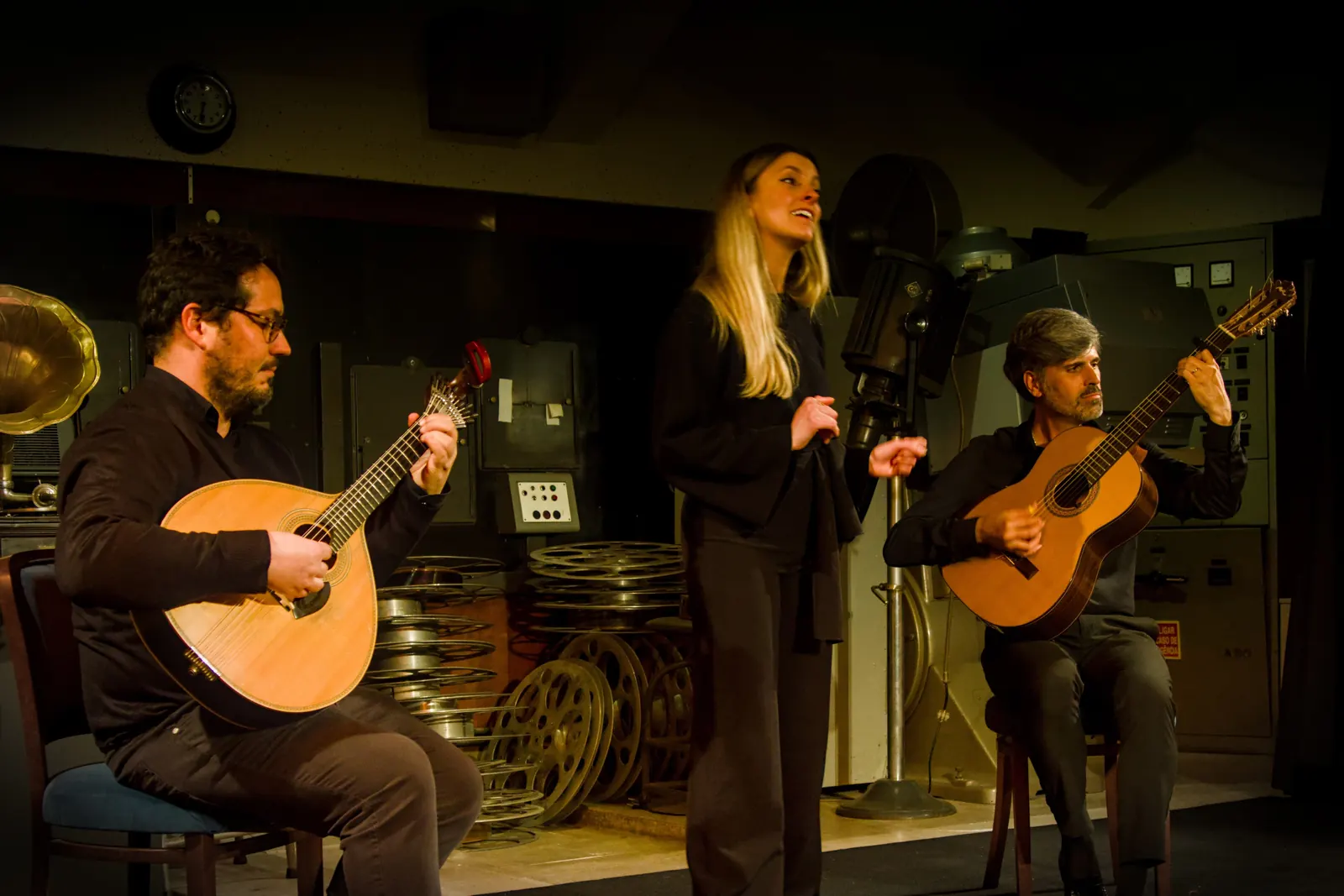 Śpiewaczka fado między dwoma siedzącymi gitarzystami — jeden trzyma gitarę portugalską, drugi gitarę klasyczną — na niewielkiej scenie